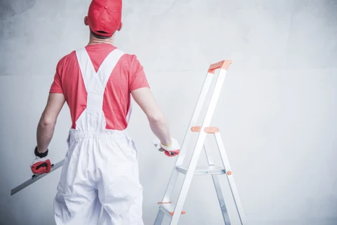 Peintre professionnel en combinaison blanche et casquette rouge tenant un pinceau, debout près d'une échelle pliante en aluminium contre un mur blanc intérieur en cours de peinture