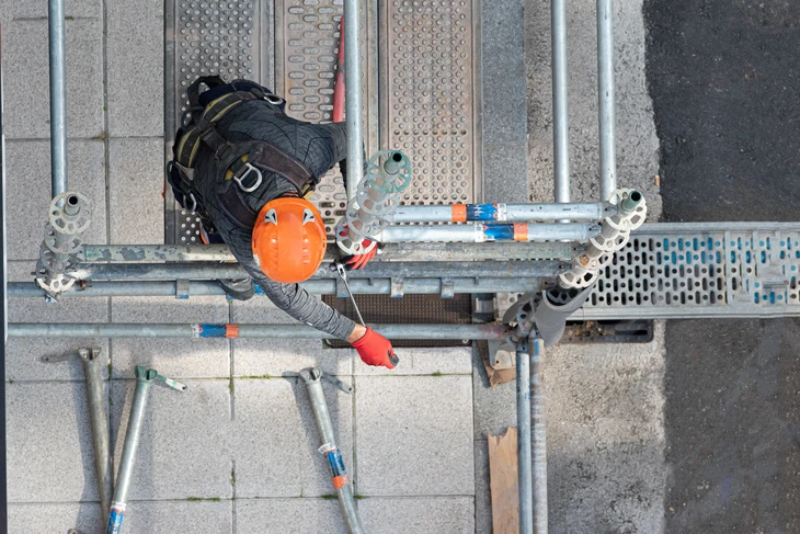 Ouvrier en hauteur sur plateforme d'échafaudage métallique, portant casque orange et harnais de sécurité, effectuant des travaux de maintenance ou construction sur structure industrielle