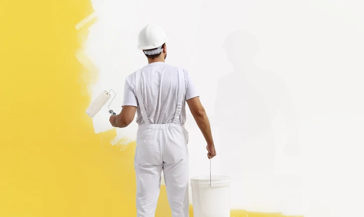 Peintre professionnel en salopette et casquette blanches appliquant de la peinture jaune sur un mur intérieur avec un rouleau à peinture