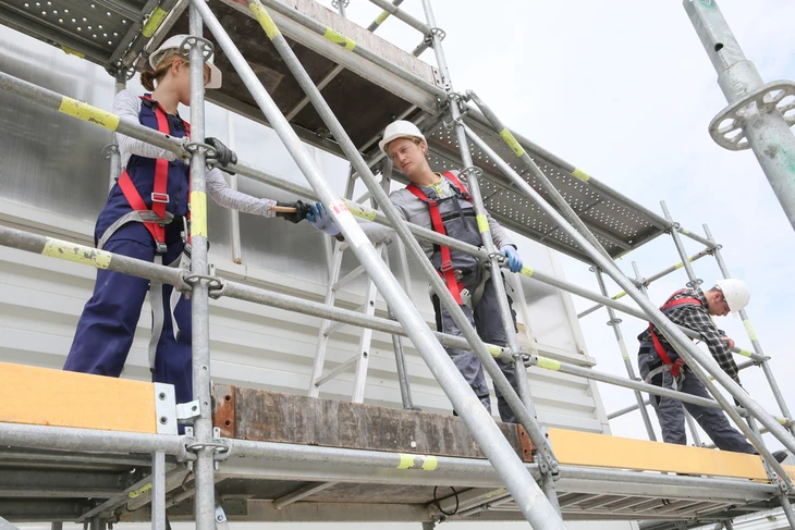 Chantier extérieur avec trois ouvriers équipés de harnais de sécurité rouge travaillant sur échafaudage métallique