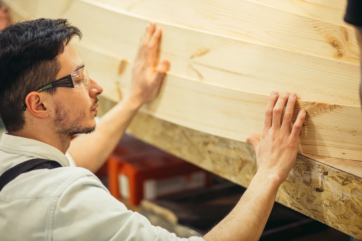 Homme portant lunettes de sécurité et chemise blanche installant une planche en bois massif contre un mur, ses mains positionnées pour ajuster l'élément
