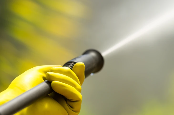 Scène extérieure de nettoyage professionnel avec pulvérisateur haute pression en action, jet d'eau visible, main gantée jaune tenant la lance de pulvérisation