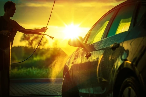 Homme nettoyant sa voiture au jet d'eau haute pression dans une allée résidentielle au coucher du soleil