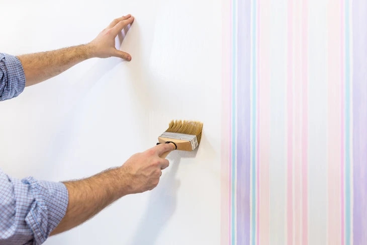 Un homme applique de la colle murale avec un pinceau large sur un mur blanc, préparant la pose de papier peint à motifs rayés pastel