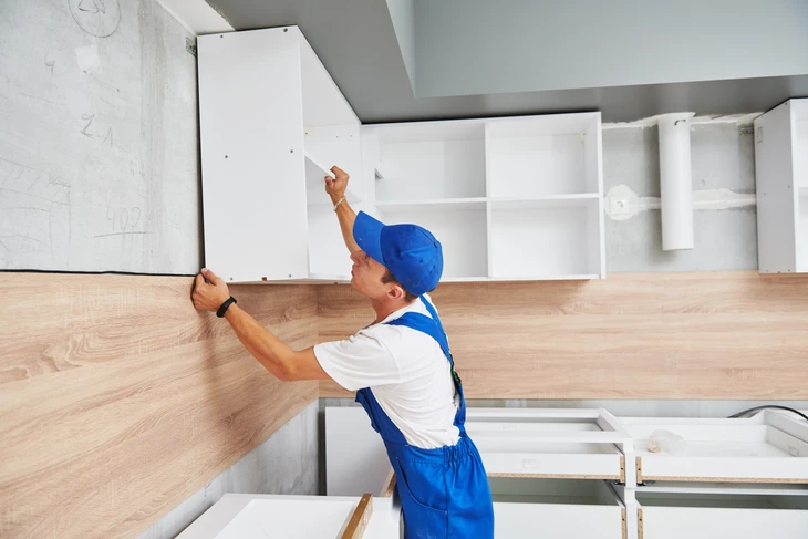 Un artisan en tenue de travail bleue et blanche avec casque installe une crédence en bois dans une cuisine moderne
