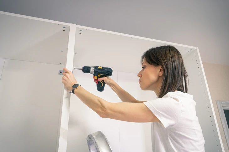Une femme en tenue blanche assemble des meubles de cuisine dans un intérieur moderne, utilisant une perceuse-visseuse sans fil pour fixer les éléments muraux blancs