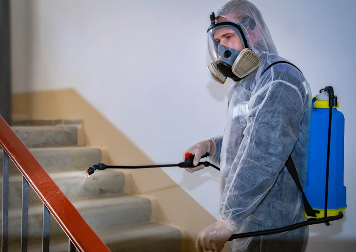 Intérieur résidentiel avec escalier, technicien en combinaison de protection intégrale et masque respiratoire effectuant pulvérisation désinfectante