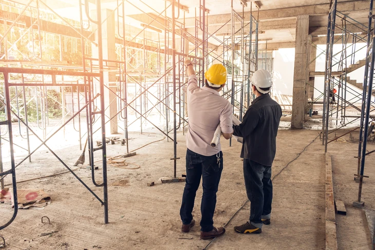 Deux hommes en casques de chantier jaune et blanc inspectent un bâtiment en construction, entourés d'échafaudages métalliques