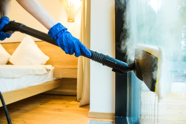 Personne en gants bleus utilisant un nettoyeur vapeur professionnel dans un intérieur résidentiel avec escalier en bois et baie vitrée
