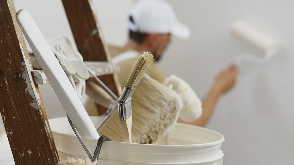 Personne en combinaison bleue de travail appliquant de la peinture blanche sur un mur intérieur avec un pinceau plat