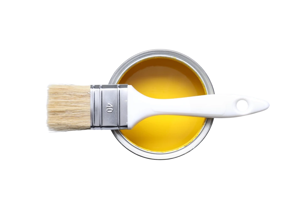 Scène d'intérieur montrant un pinceau plat à manche blanc et virole dorée trempé dans un pot de peinture rouge bordeaux, posé sur des planches de bois peintes en rouge et blanc avec finitions patinées