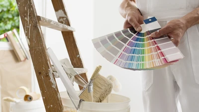 Homme souriant en salopette blanche et chemise à carreaux tenant un nuancier de couleurs et un rouleau à peinture dans un intérieur lumineux avec escabeau en bois
