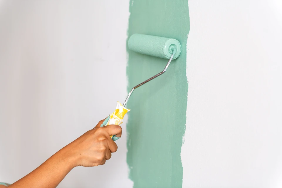 Photographie réelle montrant un artisan peintre professionnel en train de préparer ou de peindre un mur intérieur. L'image doit capturer le travail actif : application de peinture au rouleau ou à la brosse sur un mur, préparation des surfaces (ponçage, enduit), ou finitions soignées. L'artisan doit être équipé professionnellement (combinaison, protections) dans un intérieur en cours de rénovation. L'accent doit être mis sur la rigueur du travail et la qualité de l'exécution.